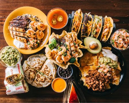 An overhead spread — tacos, a quesadilla, nachos, guacamole, shrimp and a burrito on a wood table